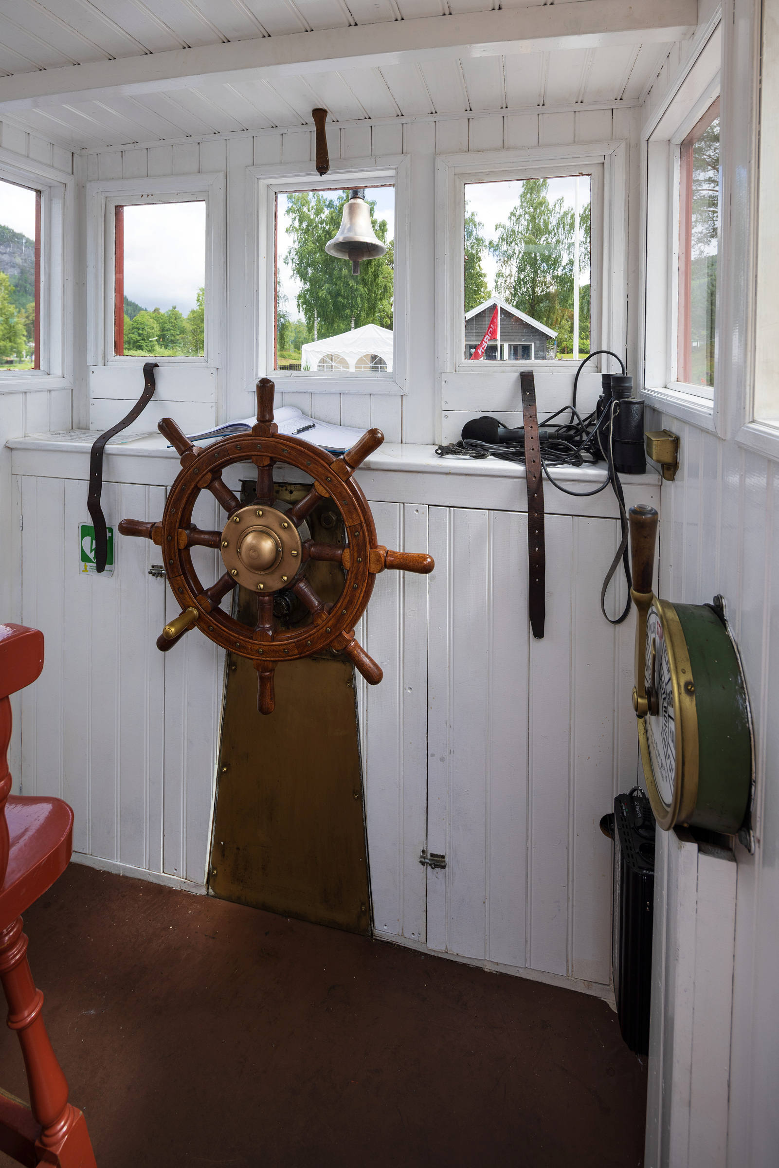 No blir D/S «Bjoren» freda 4 Skipsrattet på D/S Bjoren, og navigasjonsinstrumenter inne i den hvitmalte kahytten, med vinduer som viser trær og en rød bygning utenfor.