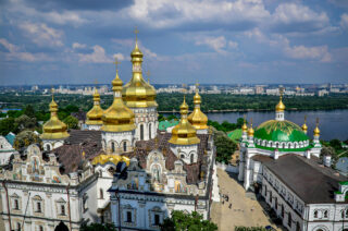 Luftfoto av klosterkomplekset Kyiv-Pechersk Lavra med gyldne kupler, hvite vegger og omkringliggende bygninger ved en elv under en delvis overskyet himmel.