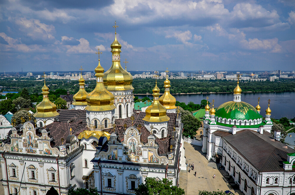 Luftfoto av klosterkomplekset Kyiv-Pechersk Lavra med gyldne kupler, hvite vegger og omkringliggende bygninger ved en elv under en delvis overskyet himmel.
