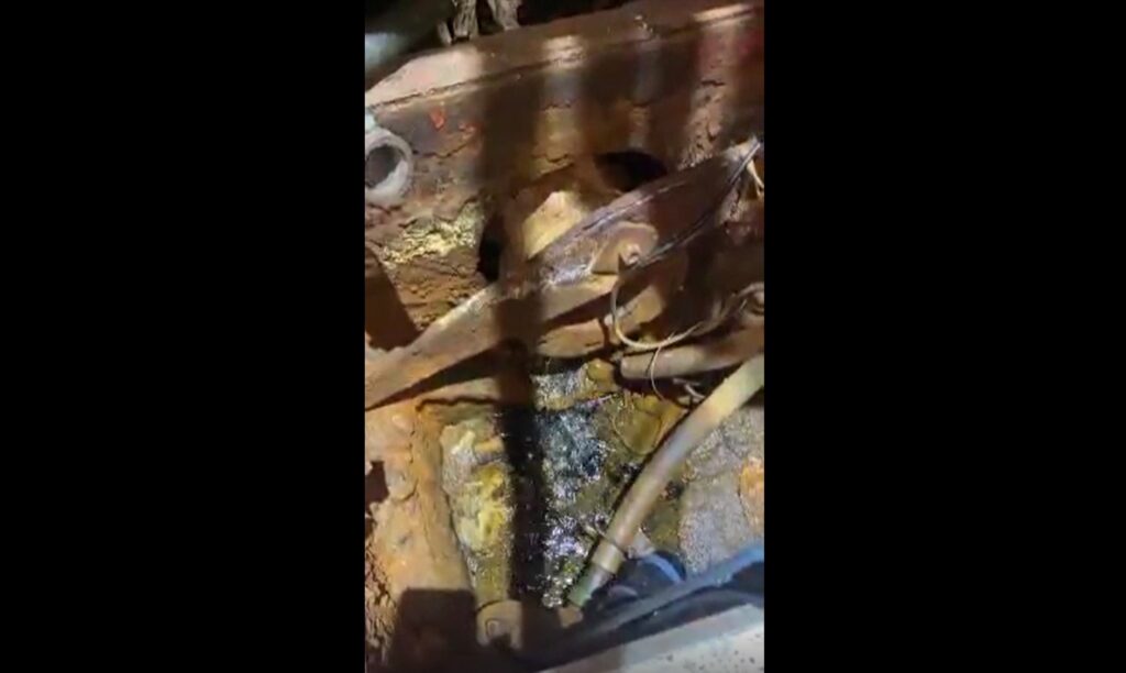 Rustne maskiner med metalldeler, slanger og olje eller væske som har samlet seg på bunnen, delvis opplyst av takbelysningen.