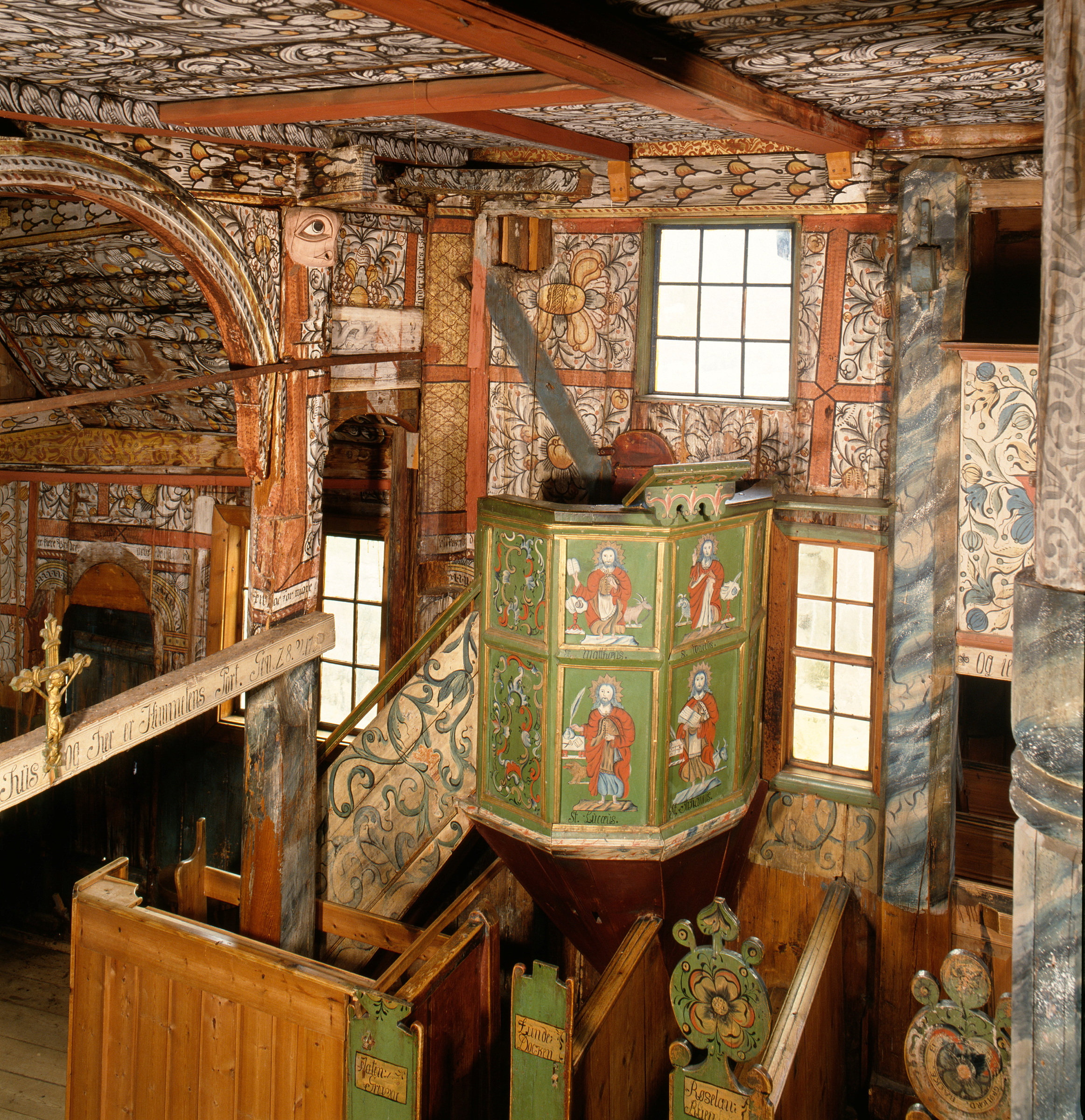 Håndtering av kirkekunst 1 Et kunstferdig malt trekirkeinteriør med en sentral grønn prekestol dekorert med religiøse figurer, omgitt av benker, utskårne detaljer og vinduer som slipper inn naturlig lys.