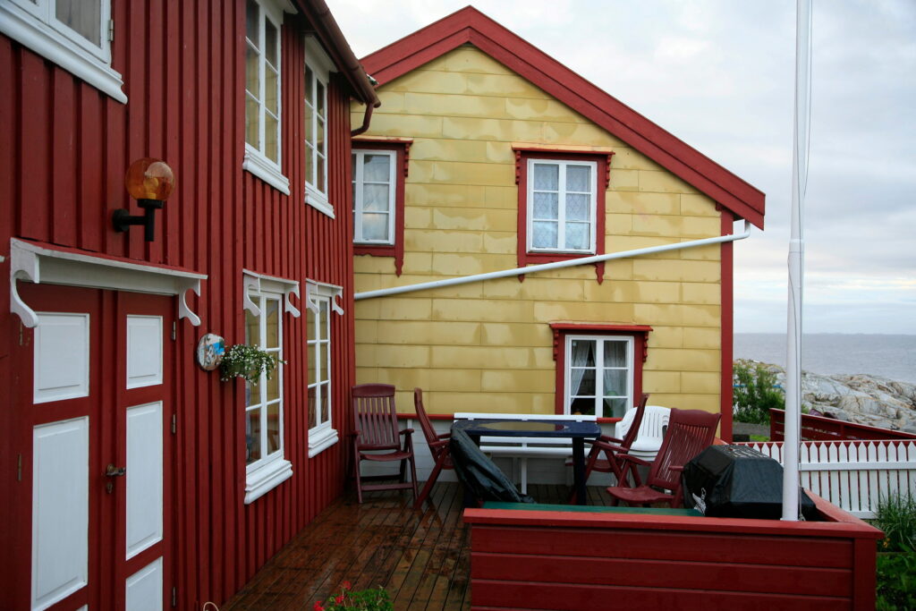 Vedlikehold av eternitt på freda og verneverdige bygninger 2 En treterrasse med utemøbler ligger mellom en rød bygning og en gul bygning, med utsikt over havet i bakgrunnen.