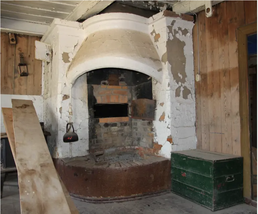 En stor, gammel, hvitkalket mursteinspeis med en buet hette, avskallet maling og synlige murstein, som står inntil vegger kledd med trepaneler; en grønn kiste og en treplank står i nærheten.