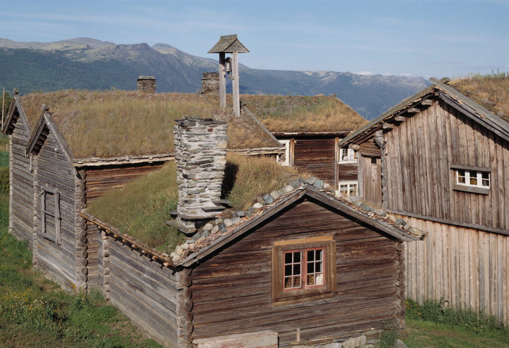En gruppe rustikke trehus med gresstak, omgitt av et fjellandskap under en klar himmel.