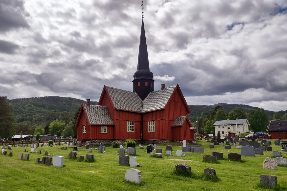 Når ein lyt jekke seg opp litt 5 En rød trekirke med et høyt tårn står på en gresskledd kirkegård med gravsteiner, med trær og en overskyet himmel i bakgrunnen.