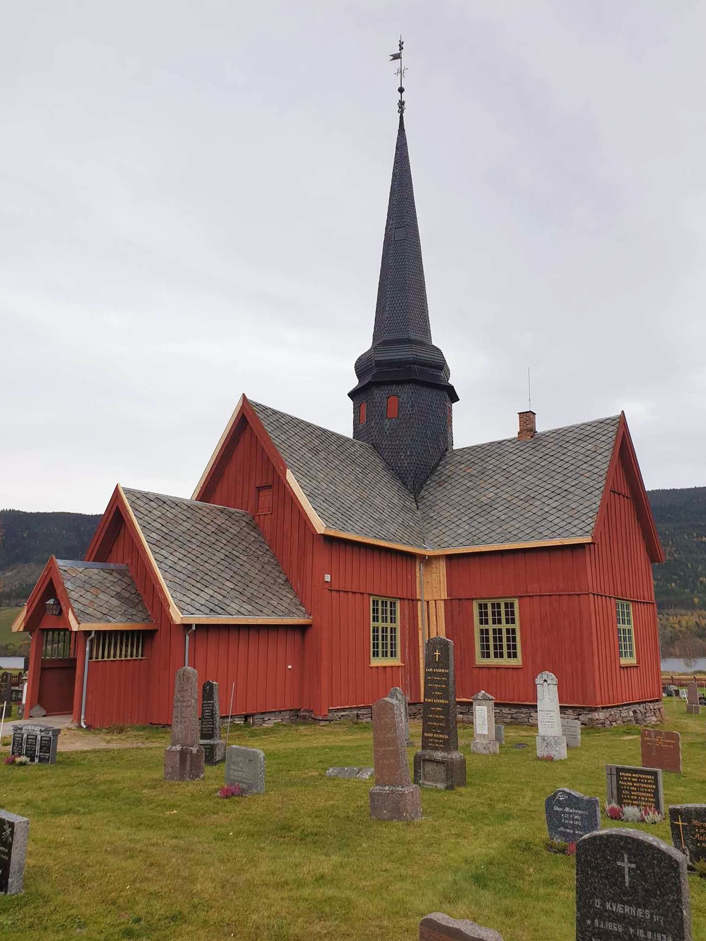 Når ein lyt jekke seg opp litt 26 En rød trekirke med et høyt, mørkt tårn, omgitt av gravsteiner på en gresskledd kirkegård, med åser og en skyet himmel i bakgrunnen.