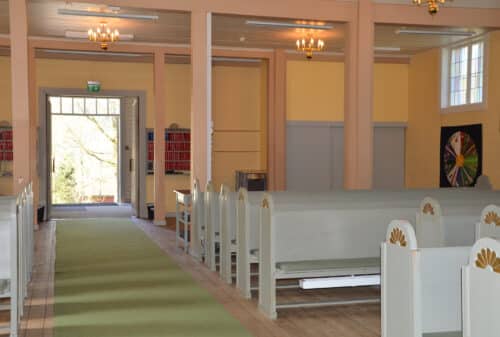 Interiøret i en kirke med hvite trebenker, en midtgang med grønt teppe, lysekroner og en dør som vender ut mot utsiden i bakgrunnen.
