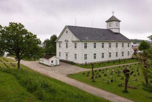 En stor, hvit trekirke med klokketårn, omgitt av en kirkegård med gravsteiner og grønne plener under en overskyet himmel.