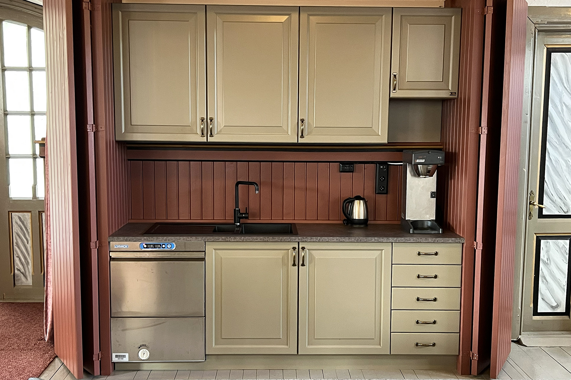 Et kompakt kjøkken med beige skap, svart vask og kran, oppvaskmaskin, vannkoker, kaffemaskin og skuffer, med en brun panelvegg i bakgrunnen.