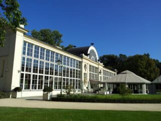 En stor bygning med glassfasade, høye vinduer, et sentralt buet vindu og tilstøtende paviljonglignende bygninger, som ligger i en anlagt hage under en klar blå himmel.