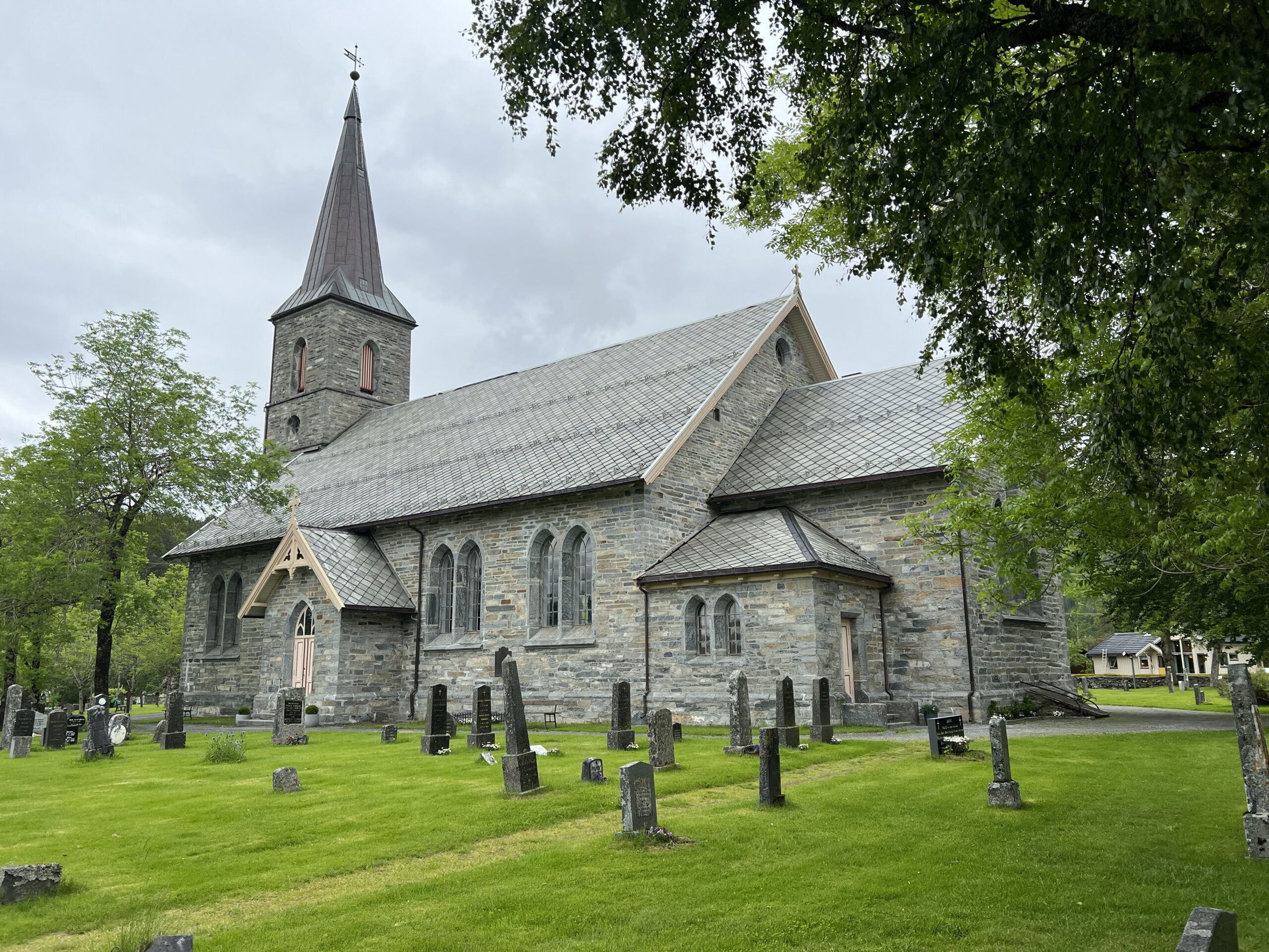 En steinkirke med et høyt tårn, omgitt av en grønn plen og gravsteiner, under en overskyet himmel.