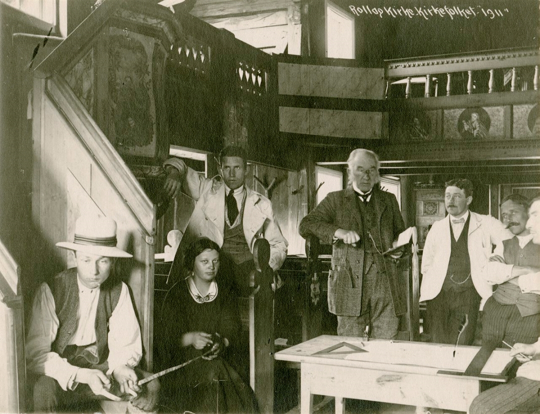 Seks personer er samlet i et rom med trepanel og religiøse malerier på veggene; to menn står, de andre sitter, og et flagg henger over dem. Bildet har tittelen «Rollag Kirke, kirkebenk 1911».
