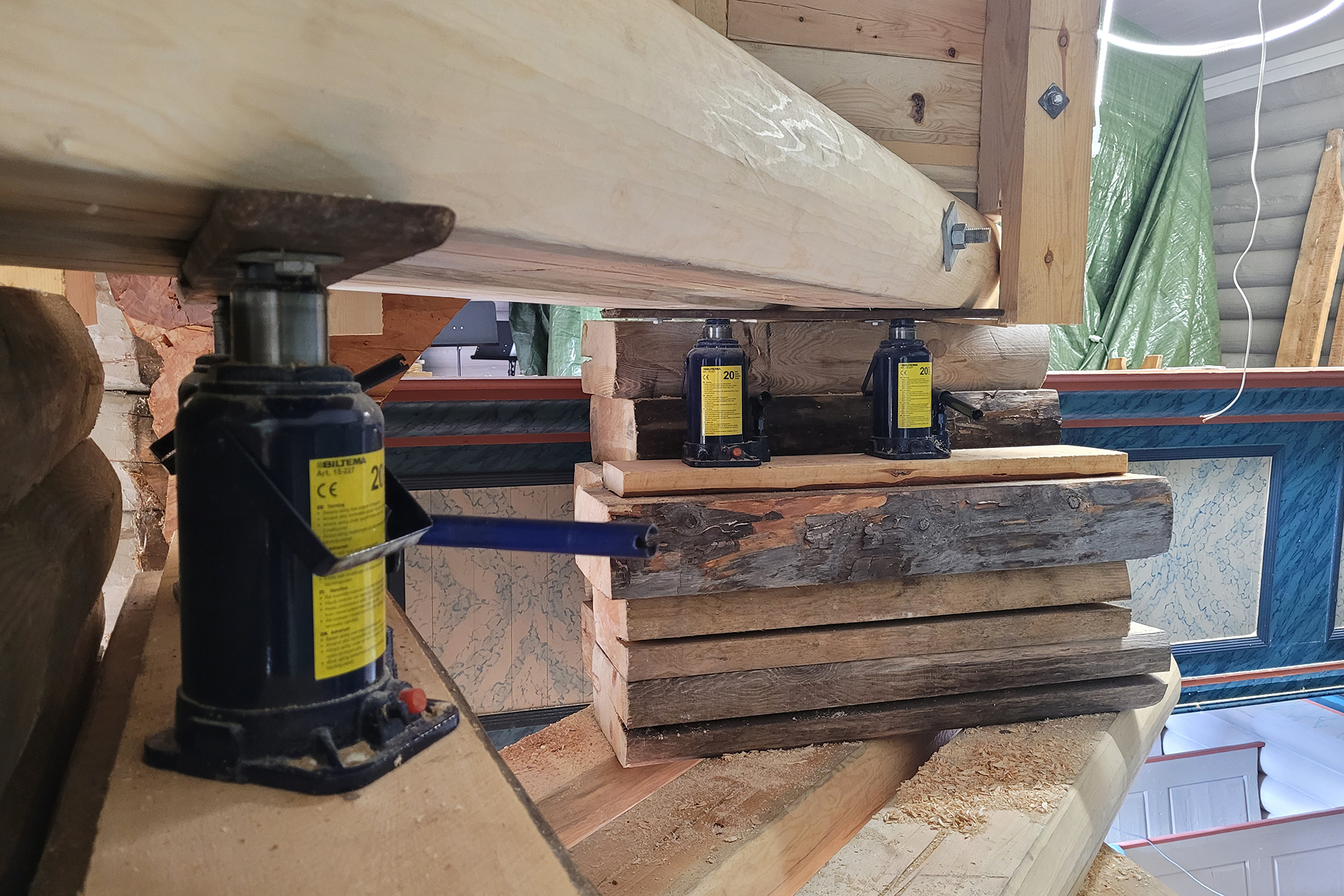 Når ein lyt jekke seg opp litt 17 To hydrauliske flaskekjekker og stablet trebjelker støtter en stor horisontal tømmerstokk inne i en bygning som er under oppføring eller renovering.