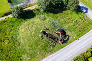 Luftfoto av et utgravningsområde ved siden av en vei, der arbeidere og maskiner graver ut et rektangulært område i en gressmark.
