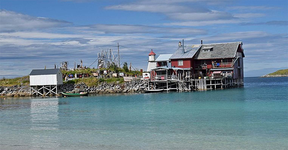 En rød trebygning og et lite hvitt skur ligger ved det klare, blå vannet, med en båt på land og et gresskledd område i bakgrunnen under en overskyet himmel.