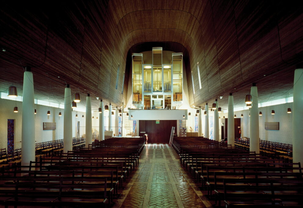 Interiøret i en moderne kirke med tretak, benkerader, flislagt gulv, hengende lamper og et stort pipeorgel foran i kirken.