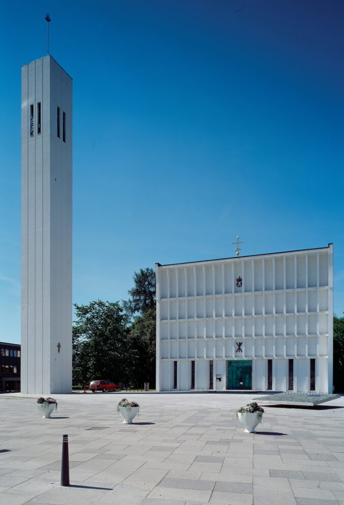En moderne hvit kirke med et høyt, rektangulært klokketårn står ved siden av en firkantet hovedbygning på et asfaltert torg under en klar, blå himmel.