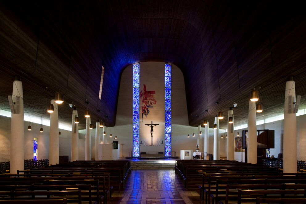 Interiøret i en moderne kirke med hvelvet tak i tre, rekker med kirkebenker, hvite søyler, glassmalerier og et krusifiks ved alteret foran.