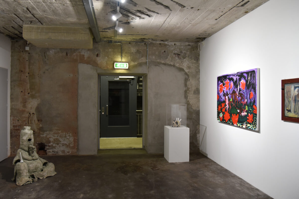 Et kunstgallerirom med betonggulv og -tak, skulpturer til venstre og i midten, to fargerike malerier på høyre vegg og en utgangsdør i bakgrunnen.