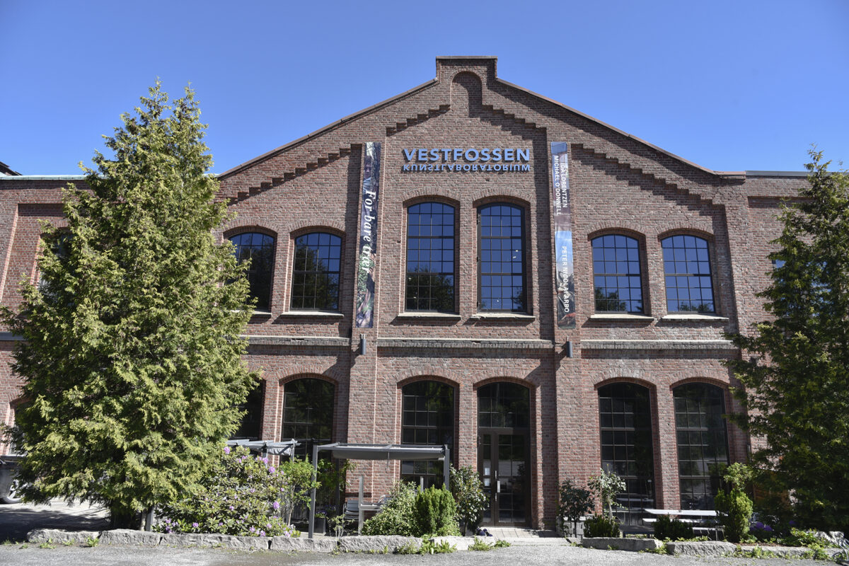 En stor murbygning med høye vinduer og skiltet «Vestfossen Kunstlaboratorium» over inngangen, omgitt av trær under en klar, blå himmel.