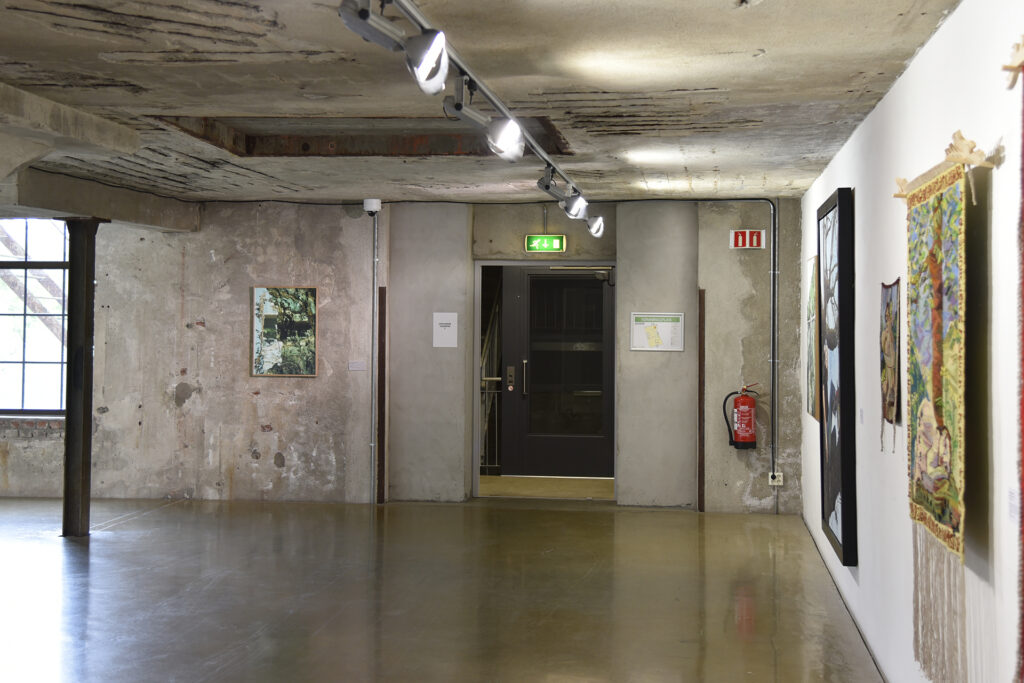 Kunstgalleri med betongvegger, utstilte malerier, nødutgangsdør i midten, brannslukker og belysning på skinner i taket.