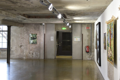 Eksempelsamling 2 Kunstgalleri med betongvegger, utstilte malerier, nødutgangsdør i midten, brannslukker og belysning på skinner i taket.