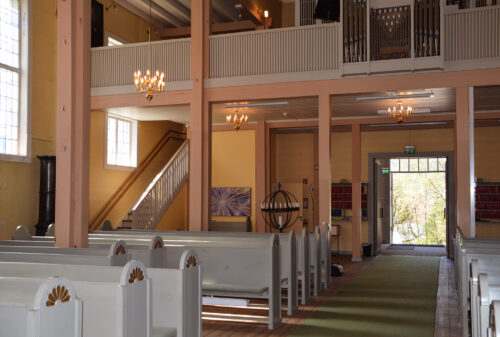 Interiøret i en kirke med hvite trebenker, gule vegger, lysekroner, en trapp og store vinduer som slipper inn naturlig lys.