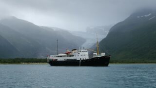 Et svart-hvitt skip flyter på rolig vann, omgitt av grønne fjell og en isbre i bakgrunnen under en overskyet himmel.