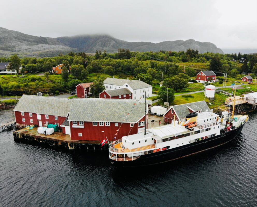 Et svart-hvitt skip lå ved kai ved siden av røde trehus langs en grønn, kupert kystlinje under en overskyet himmel.