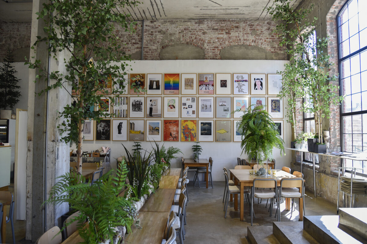 En moderne kafé med murvegger, store vinduer, mange grønne planter, trebord og en gallerivegg med fargerike, innrammede kunstverk.