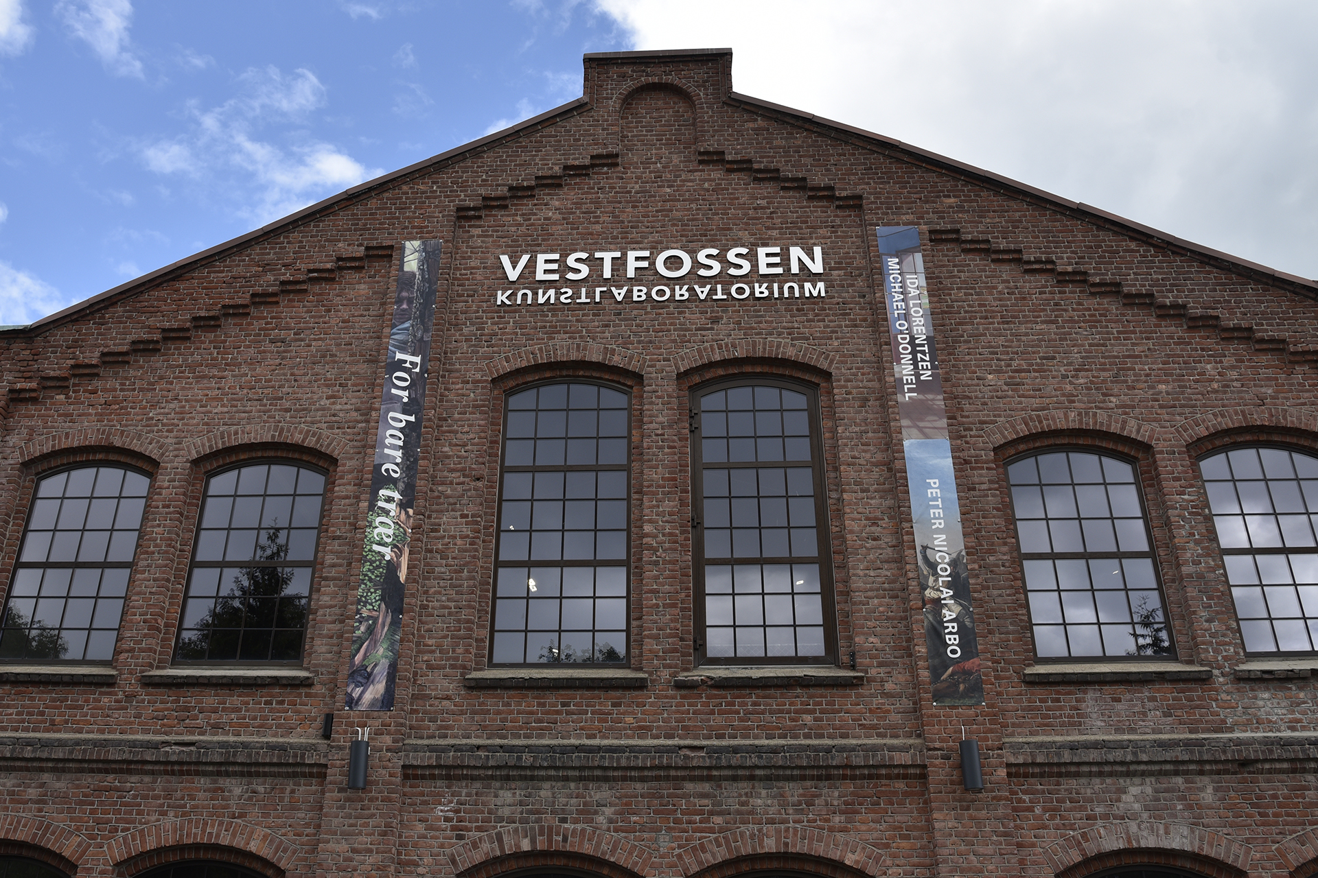 En murbygning med skiltet «Vestfossen Kunstlaboratorium» over store buede vinduer; to vertikale bannere henger på hver side av det midterste vinduet.