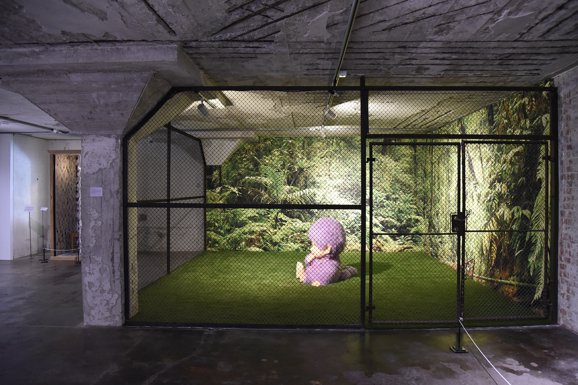 I et inngjerdet område med kunstgress sitter en rosa, tegneserieaktig figur. Veggene er dekorert med bilder av tett, grønt løvverk, noe som skaper en jungellignende bakgrunn.