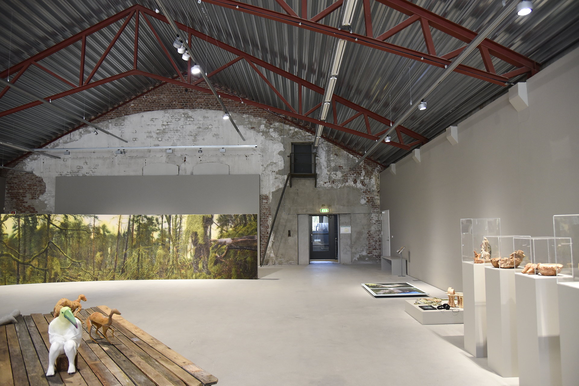 Et kunstgalleri med synlige bjelker, et veggmaleri med skogsmotiv, skulpturer på plattformer og ulike kunstverk utstilt mot nøytrale vegger.