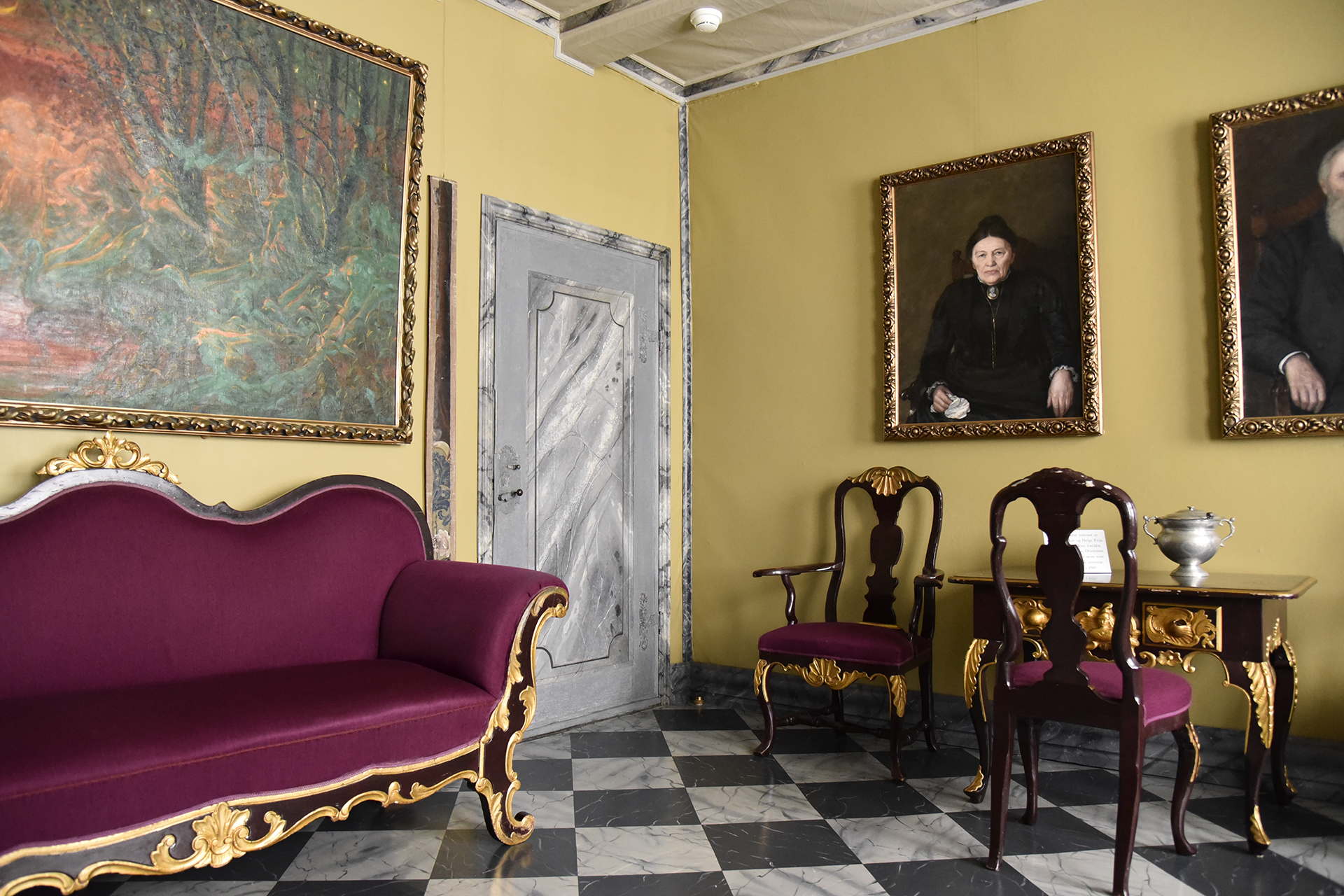 Å verne verdiane utan å skade 9 Et elegant rom med en lilla-gullfarget sofa, to stoler, et lite bord og store innrammede portretter på gule vegger, med en grå dør og et gulv med rutete fliser.