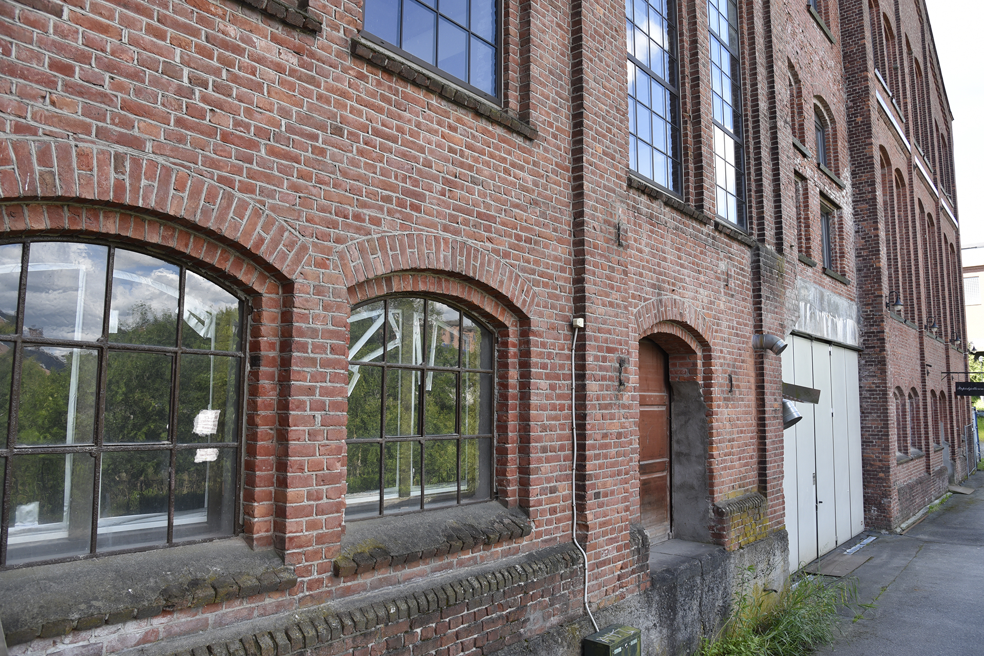 En industribygning i rød murstein med store buede vinduer, hvite dobbeltdører og en smal betongsti langs siden. Man kan se refleksjoner av trær i glasset.
