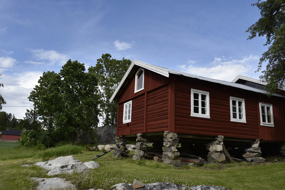 Stor istandsetting med lite endring 6 Et rødt trehus med vinduer med hvite rammer står på høye steinstøtter i en gresskledd hage med trær og steiner.