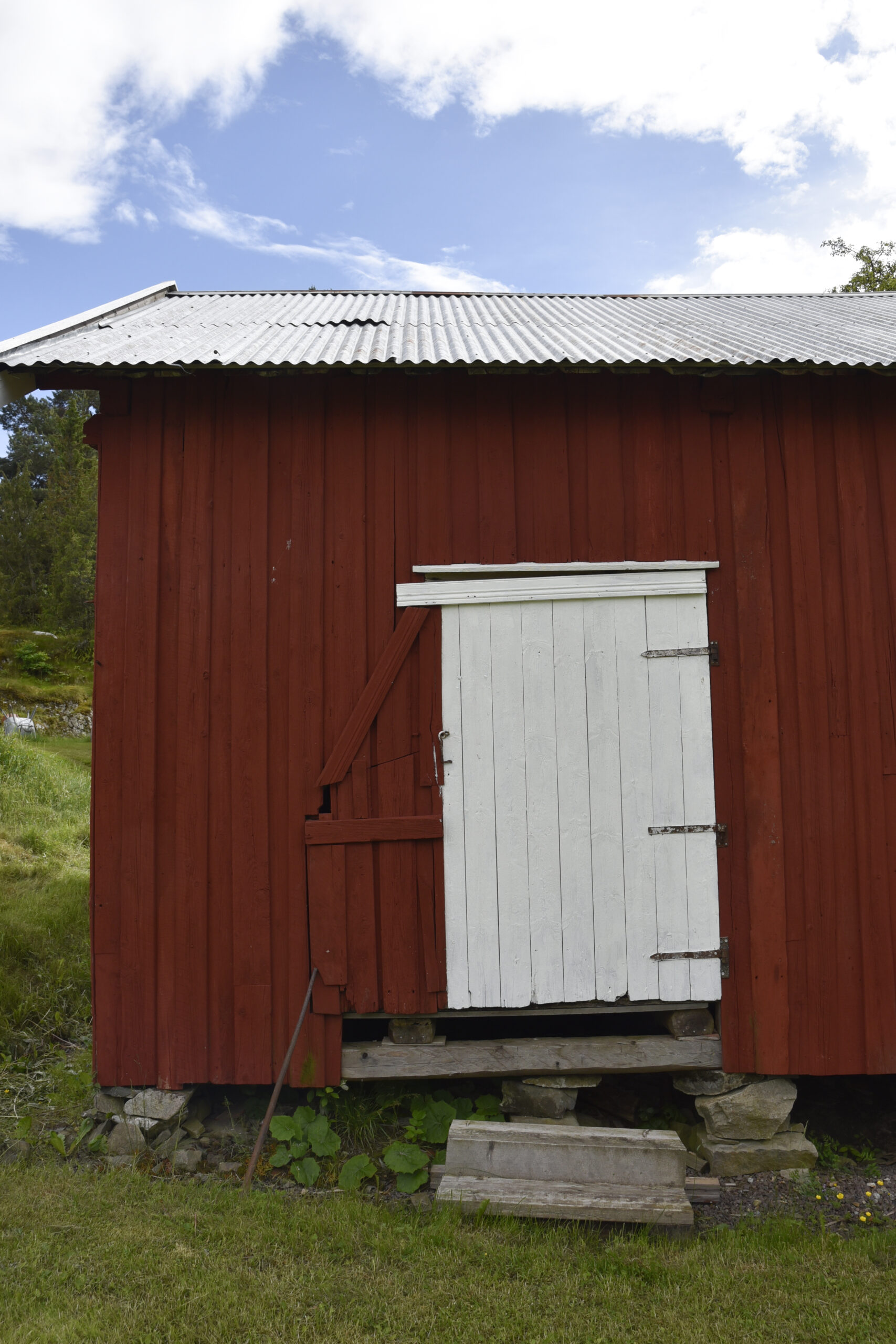 Stor istandsetting med lite endring 8 Et rødt trehus med en delvis ødelagt hvit dør står på et steinfundament under en delvis overskyet himmel.