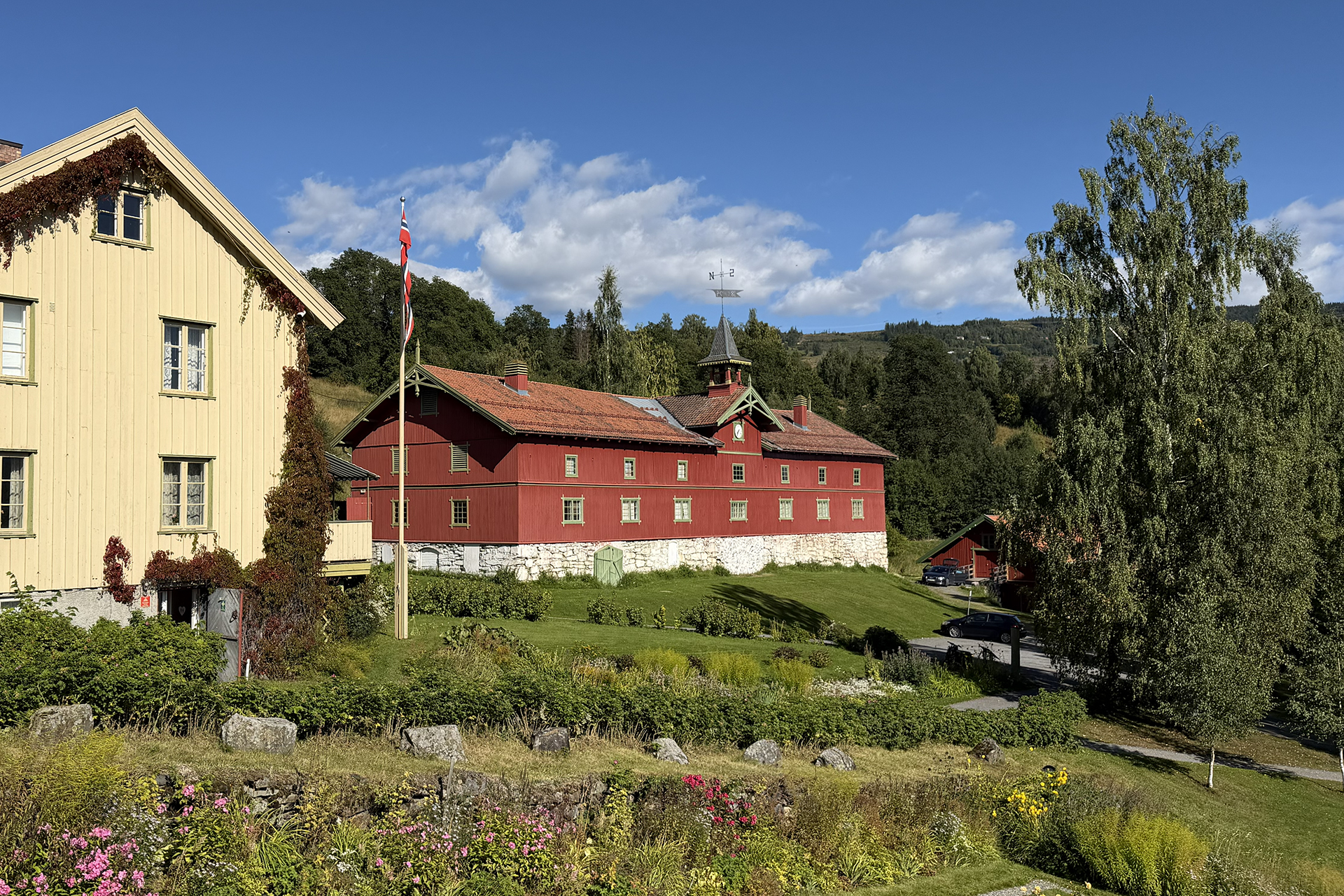 Får lov til å vere låve, men med ny bruk 6 To tradisjonelle trehus, ett gult og ett rødt, ligger på en gresskledd åsside med et norsk flagg, trær og blå himmel i bakgrunnen.