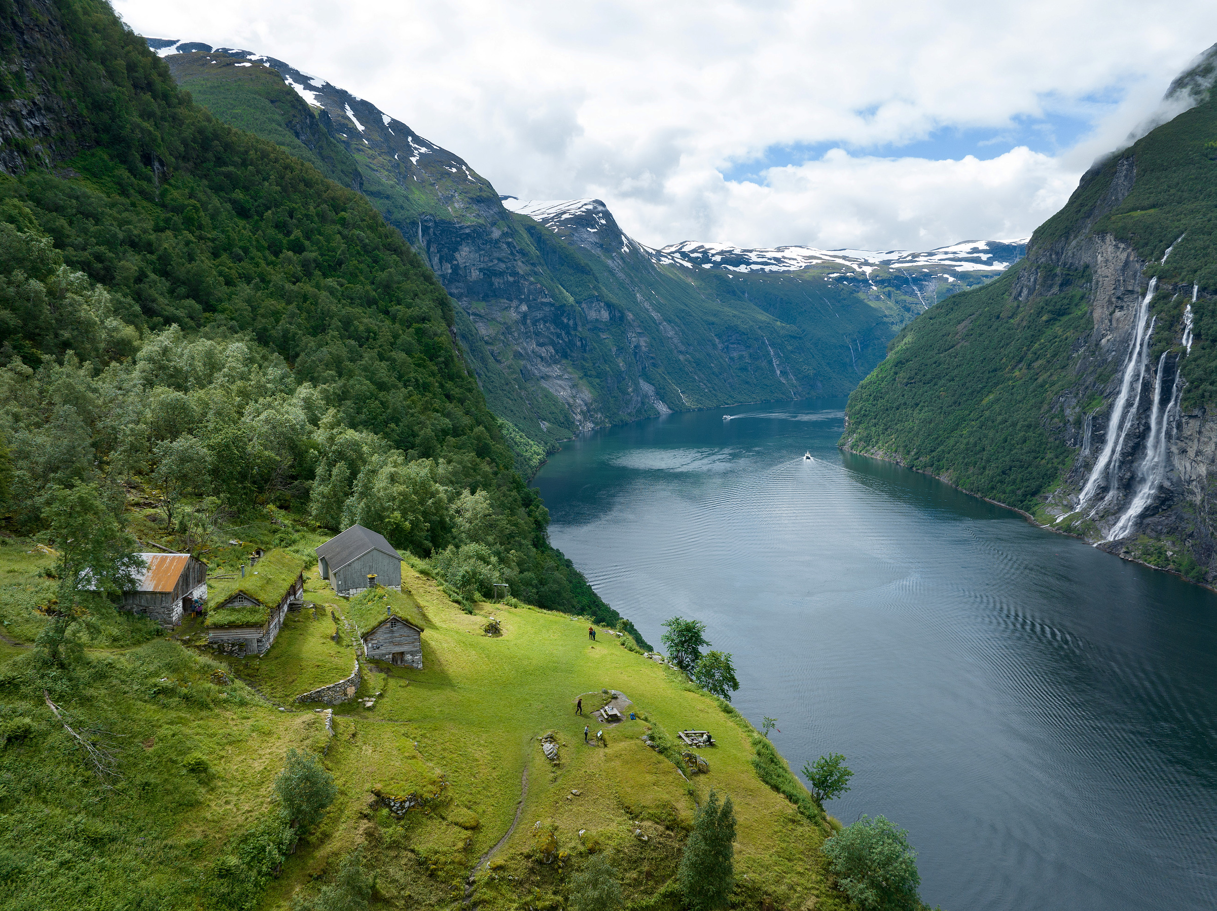 Norges verdensarv 1 En gruppe rustikke bygninger ligger på en gresskledd åsside med utsikt over en fjord – en del av Norges verdensarv – omgitt av grønne fjell og fjernliggende fosser under en skyet himmel.