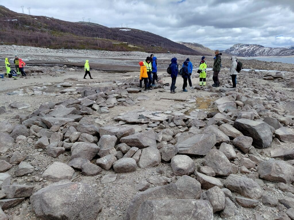 En gruppe kulturmiljøforvaltere på befaring står i friluftsklær ved tuften på Langegard ved Strandavatn, med åser i bakgrunnen.