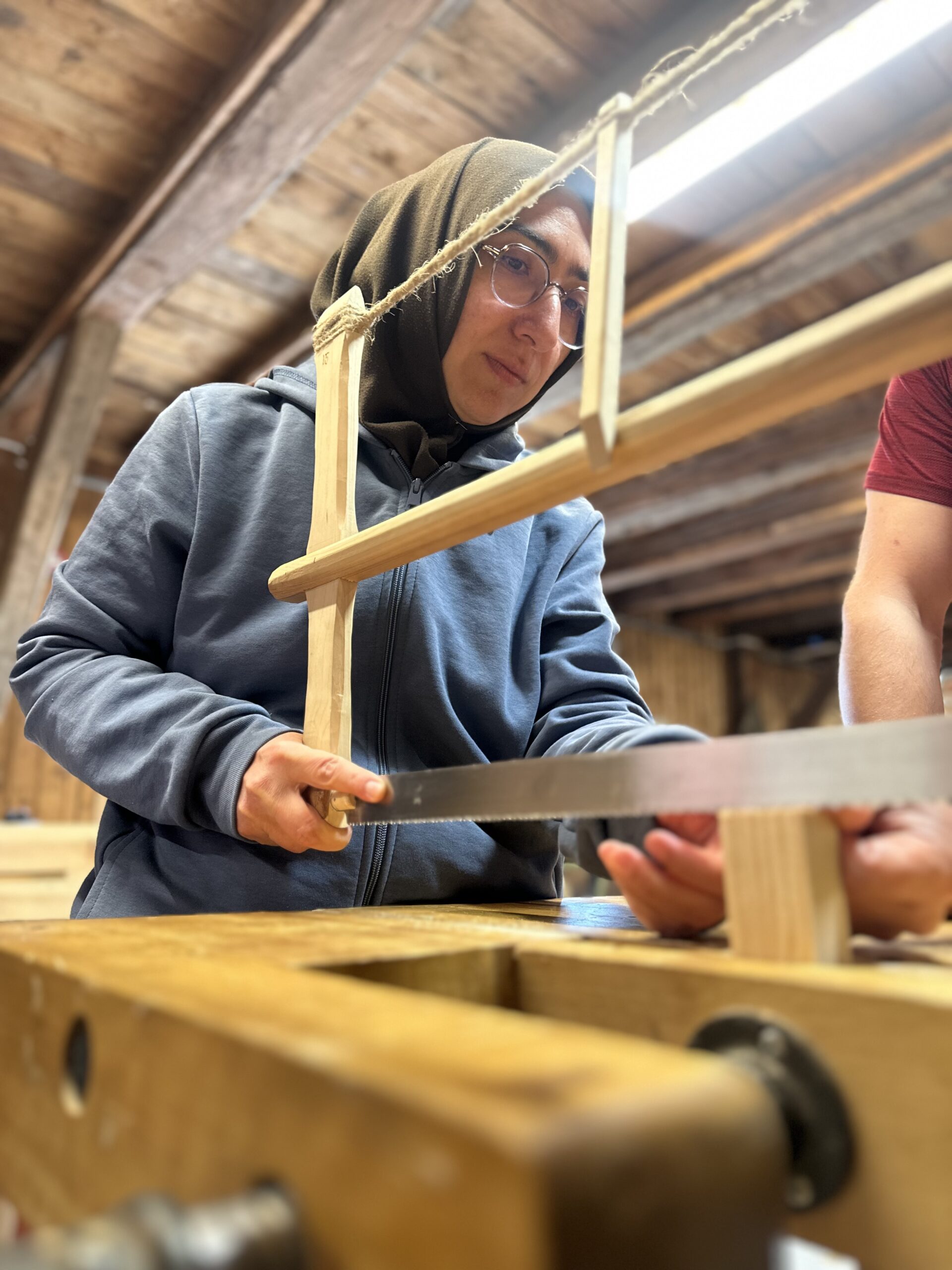 A person in a headscarf uses a wooden handsaw on a workbench, focusing intently. Another persons arm is partially visible, assisting with the task.