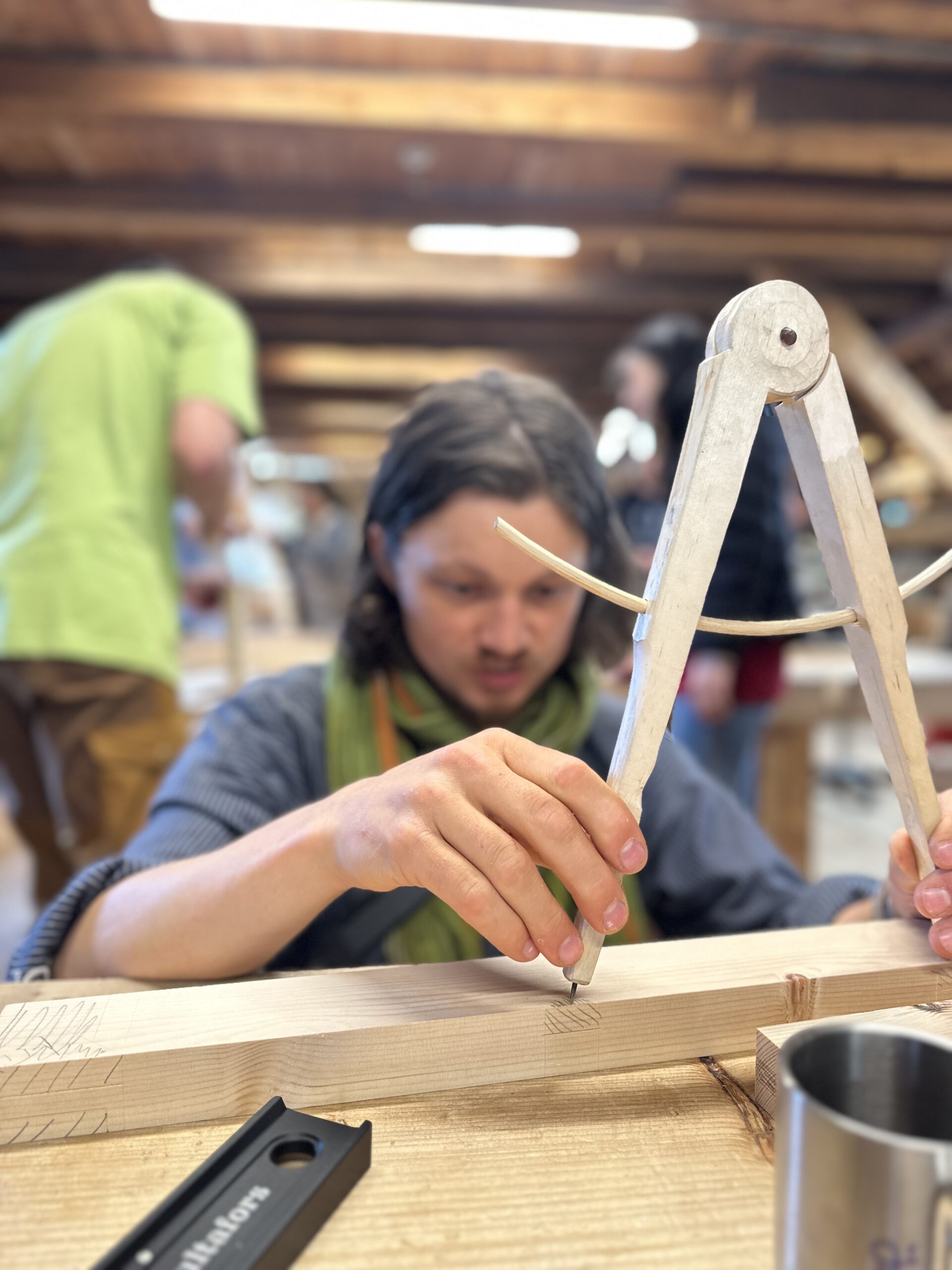 Person using a wooden compass to mark measurements on a wooden plank in a workshop, with others working in the background.