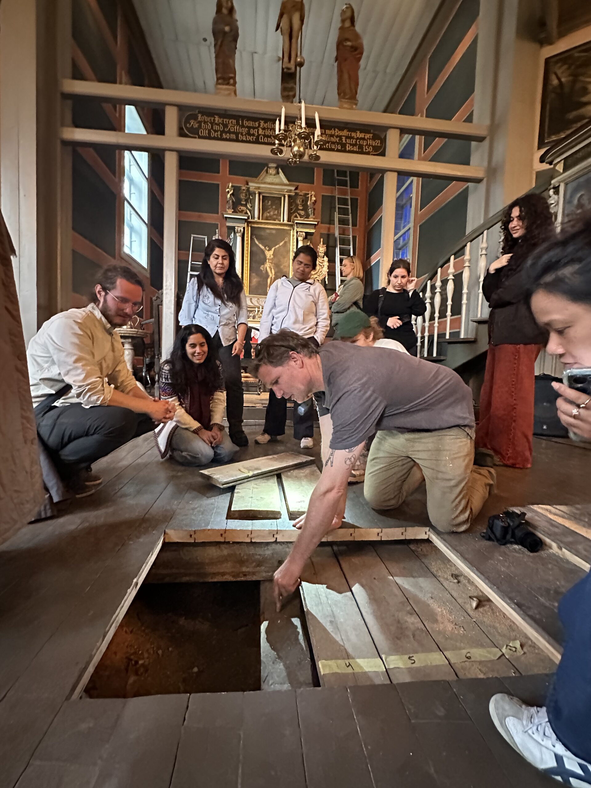 A group of people observes as a man lifts wooden floorboards to reveal a hole in the floor inside a historic building.