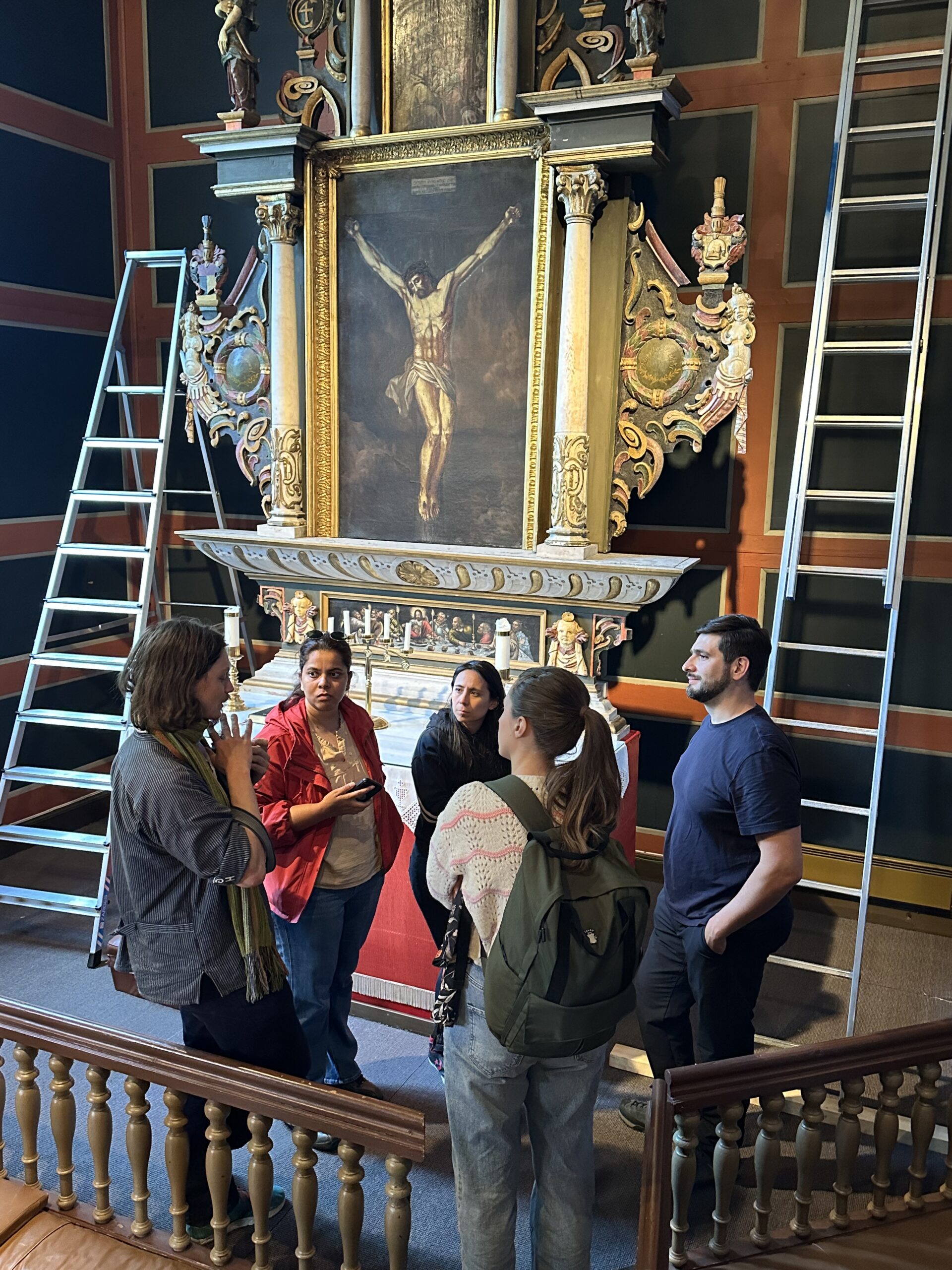 Five people stand and talk in front of an ornate crucifixion painting, with two ladders on either side, inside a dimly lit room.