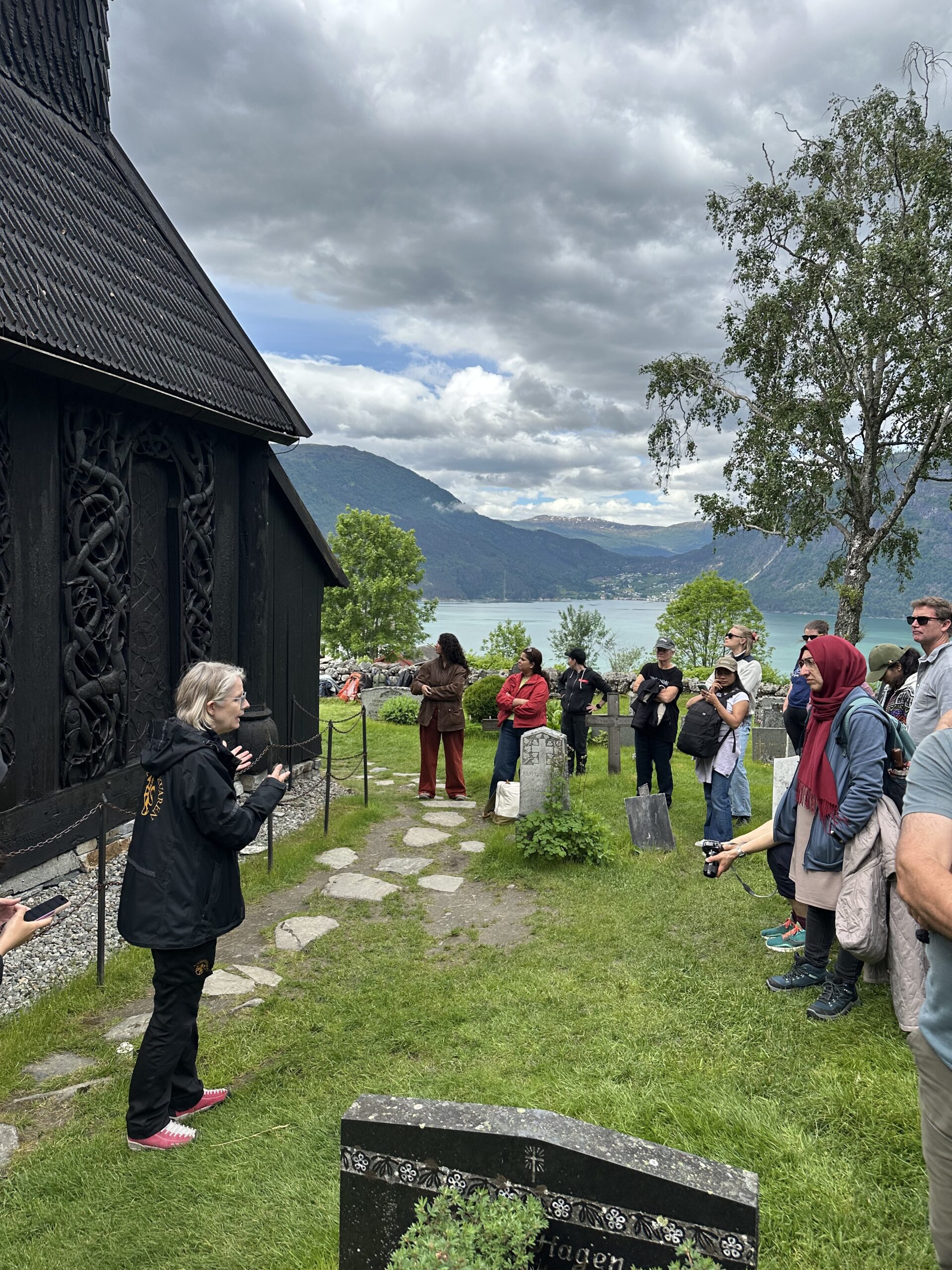 A group of tourists listens to a guide outside a dark wooden building, with mountains and a lake visible in the background under a cloudy sky.