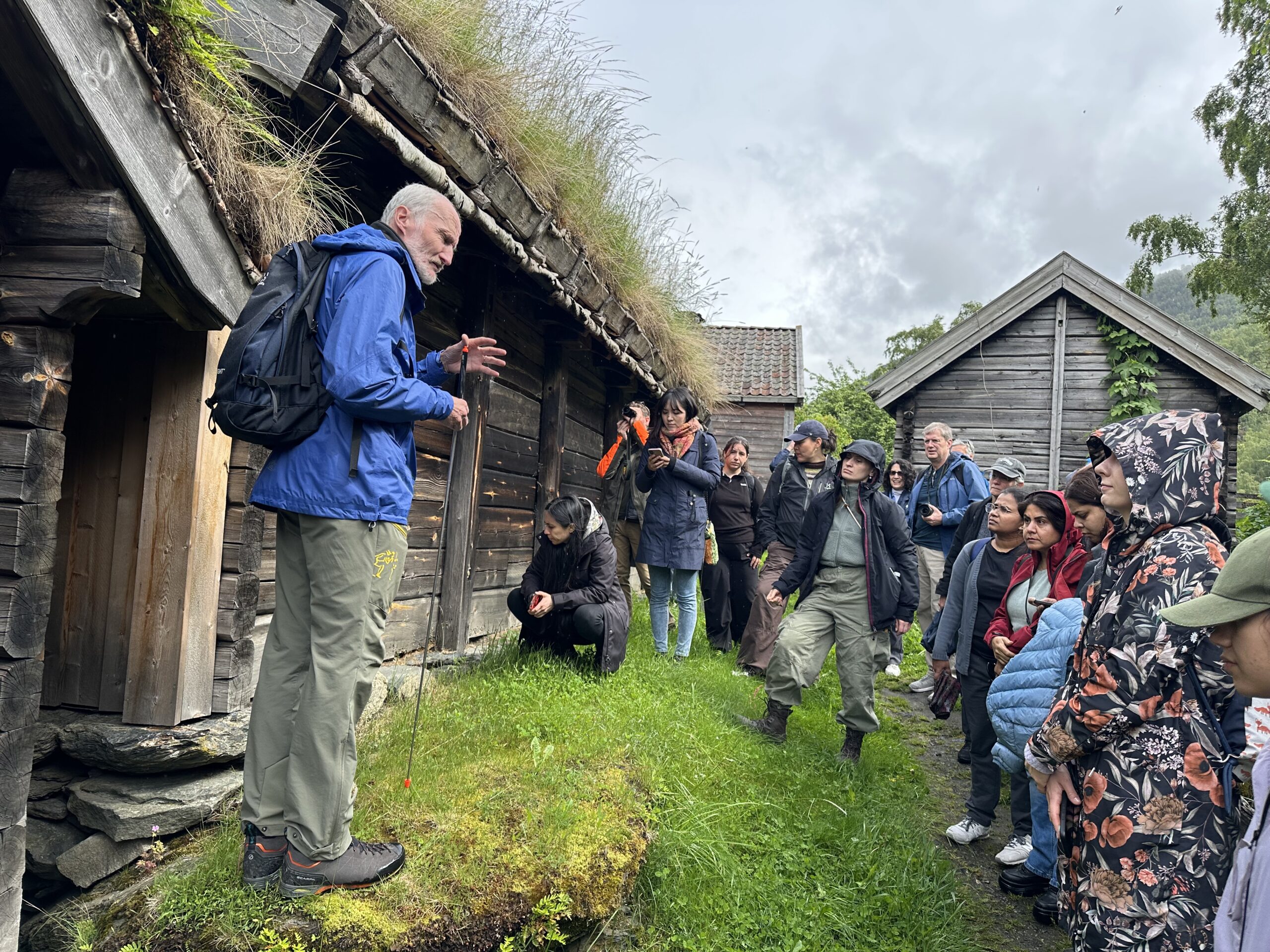 A guide in a blue jacket talks to a group of people outside old wooden houses with grass roofs on a cloudy day.