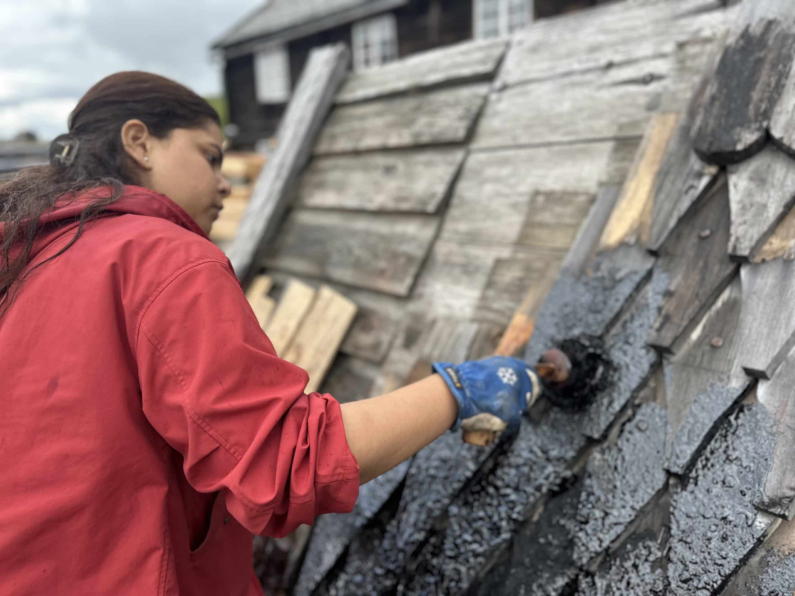 Person in a red shirt applies black sealant to wooden shingles on a sloped roof outdoors, holding a brush with a gloved hand.
