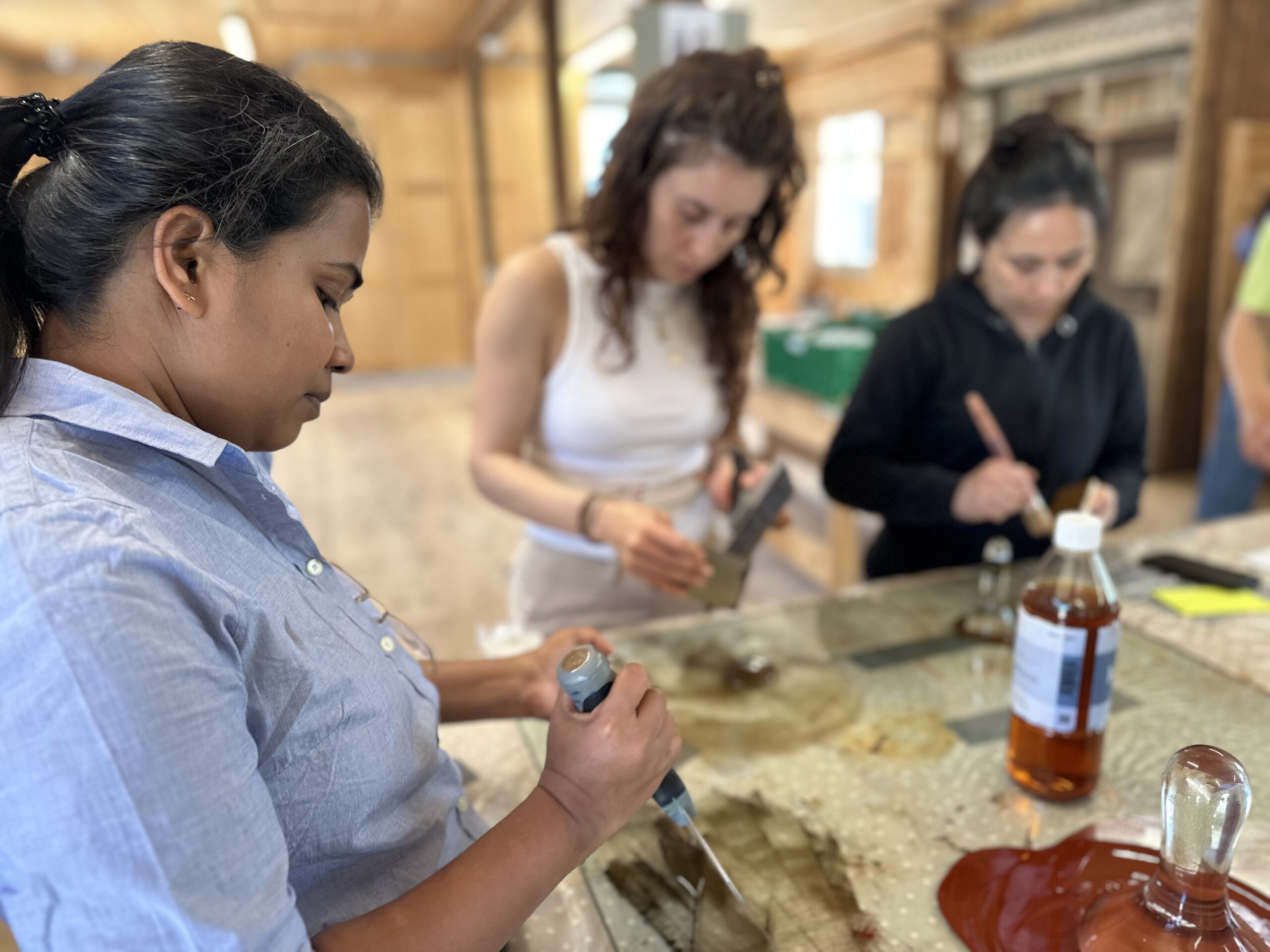 Three women stand at a table working on a science experiment with droppers, liquid samples, and glassware in a well-lit indoor setting.