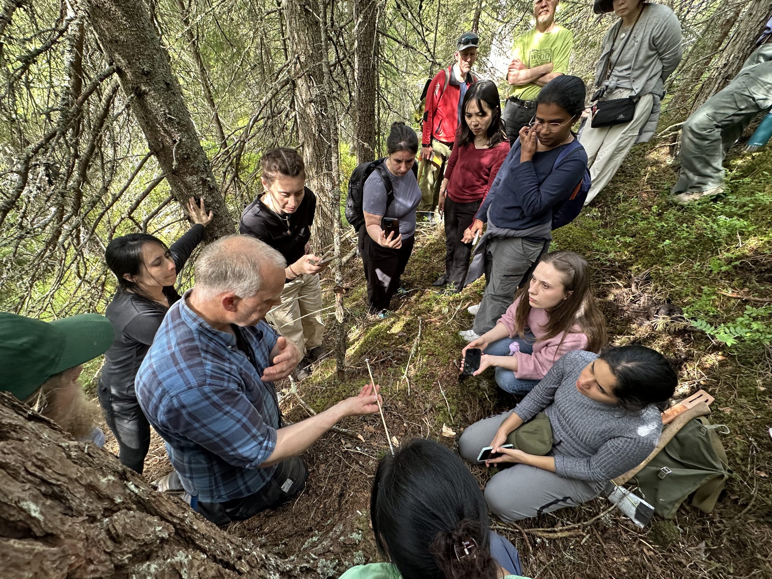 A group of people gathers in a forest while a man in a plaid shirt demonstrates something using a stick; others watch and listen attentively.