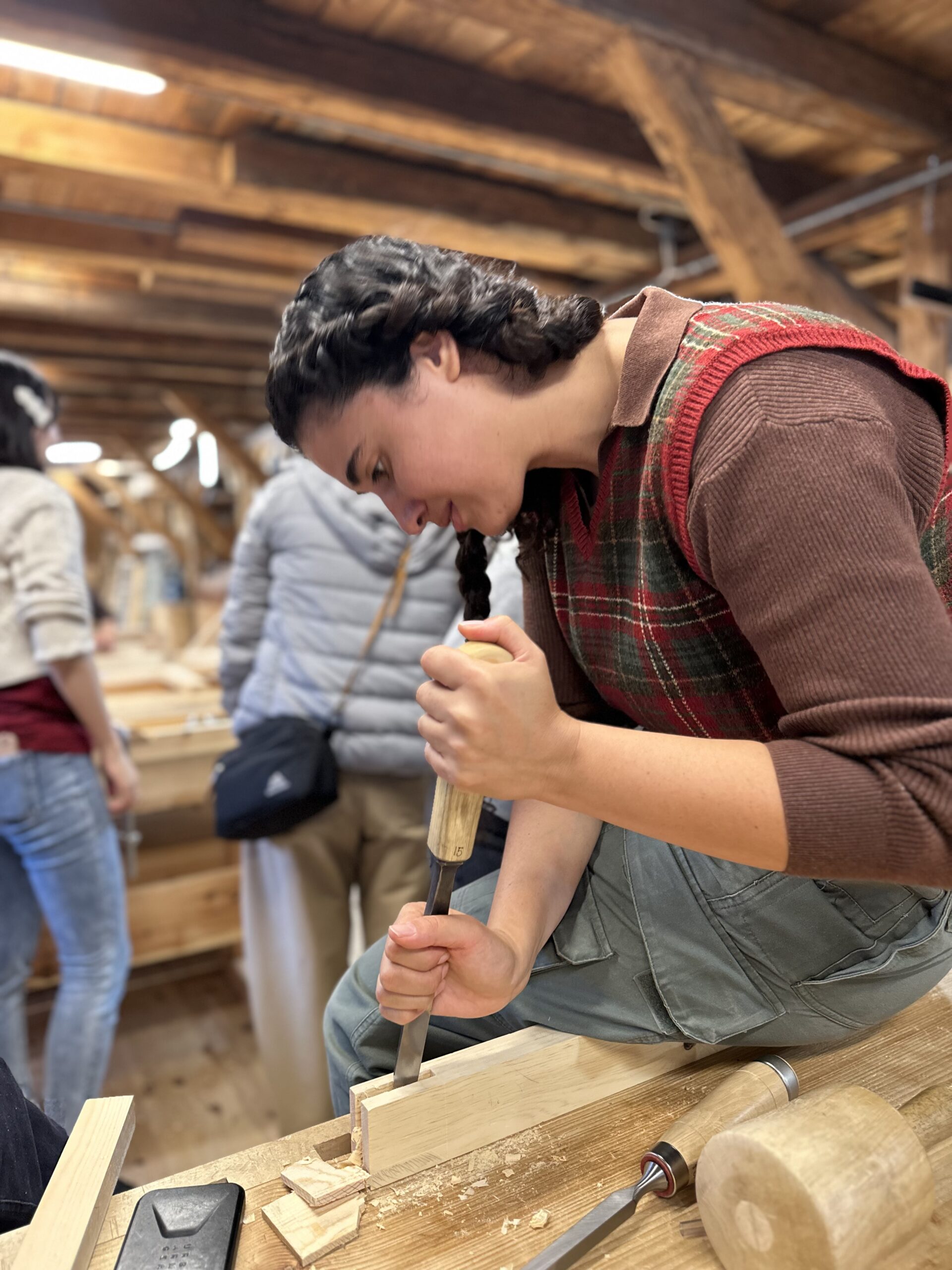 A person uses a chisel and mallet to carve a piece of wood in a workshop, with others working in the background.
